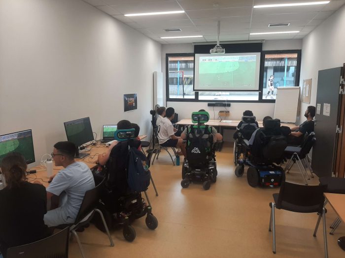 Il y a 10 personnes dans une pièce en train de jouer à des jeux vidéo de foot. Un grand écran projette l'une des parties.