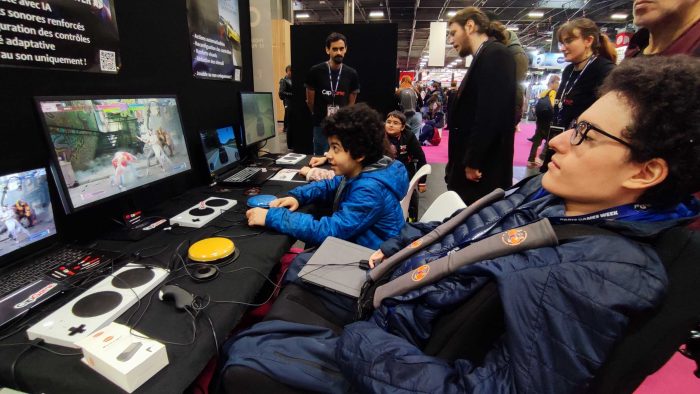 Deux frères jouent sur Street Fighter 6. Le jeune homme au premier plan, en fauteuil, ayant peu de mobilité, joue avec deux petits contacteurs pour remplacer les gros boutons du stand.