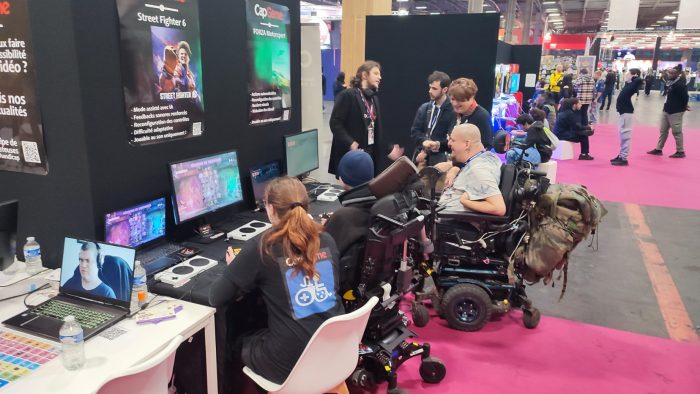 On voit le stand CapGame et ses deux postes de jeu. Les personnes sont de dos. Sur Street Fighter 6, un jeu homme en fauteuil joue avec une animatrice portant le Tshirt CapGame. Sur Forza Motorsport, un homme chauve sur fauteuil joue avec son joystick au pied.