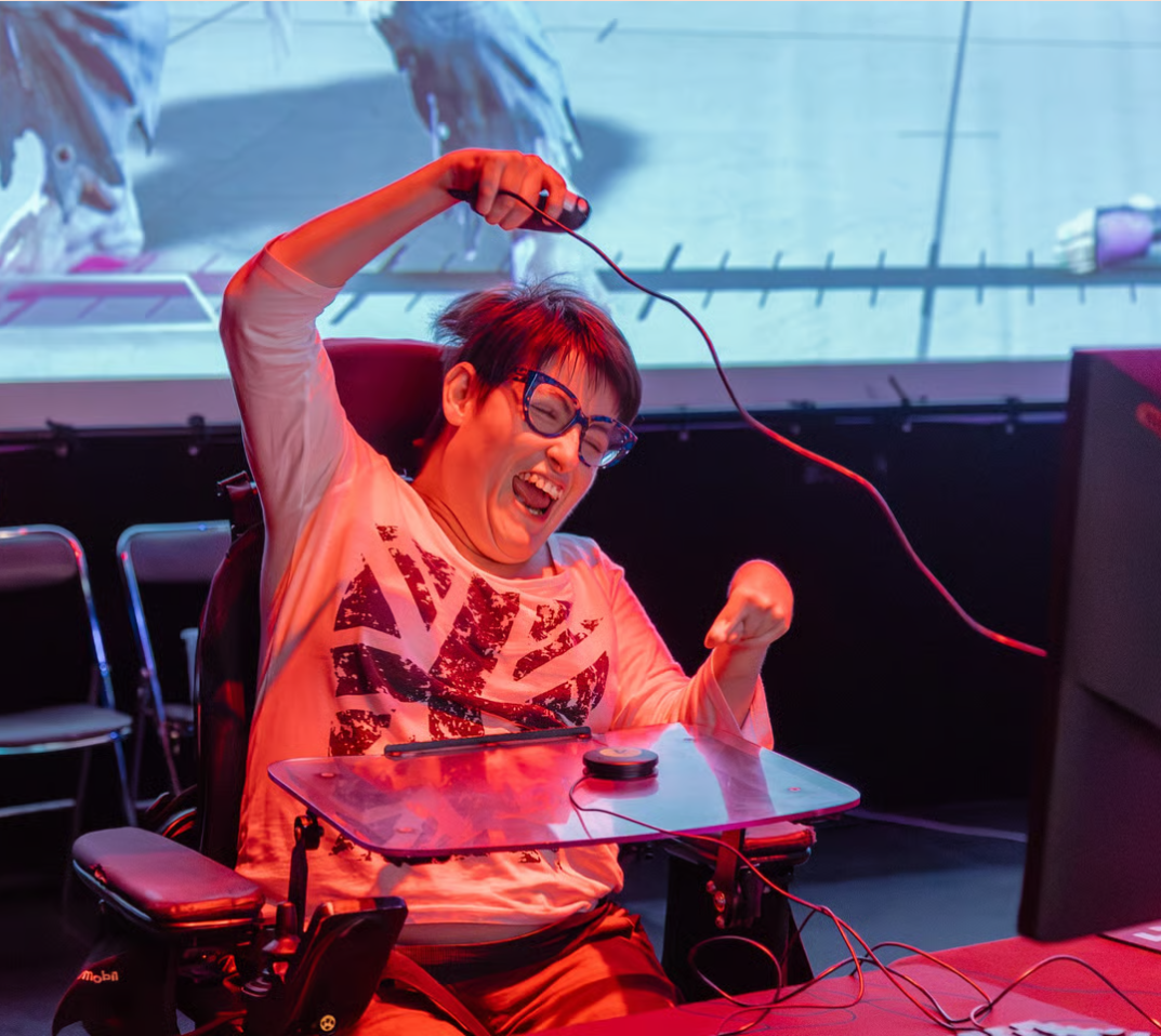 Une joueuse très joyeuse, joystick en main et un bouton sur la tablette de son fauteuil électrique. On devine Street Fighter 6 derrière elle sur écran géant.