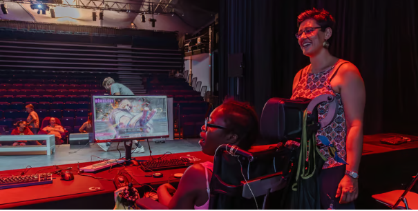La salle des congrès de Parthenay avec ses gradins en arrière plan et une jeune femme en fauteuil roulant électrique et son accompagnatrice sur la scène devant un PC faisant tourner SF6. On voit du matériel adapté sur le bureau.
